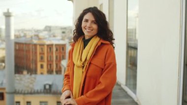 Turuncu ceketli güzel esmer kadın çatı terasında korkulukların yanında duruyor ve mutlu bir gülümsemeyle kameraya poz veriyor.