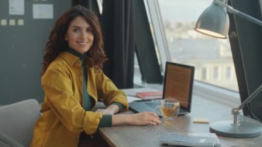 Modern ofiste masa başında oturan güzel beyaz iş kadını portresi, kameraya bakıyor ve gülümsüyor.