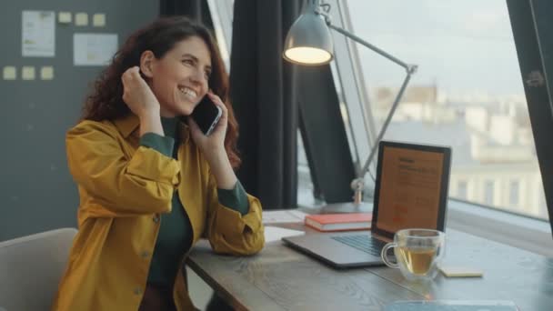 正の白人実業家女性ですカジュアルウェアで机の上に座っている現代のオフィスの窓からティーカップとラップトップで 笑顔と仕事中の携帯電話で話す — ストック動画