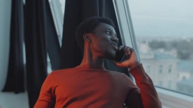 Genç Afrikalı Amerikalı adam cep telefonuyla iş tartışıyor ve modern çatı katının penceresinden bakıyor.