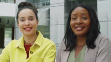 Çok ırklı iki iş kadınının portresi ofiste birlikte oturuyor, kameraya bakıyor ve gülümsüyor.