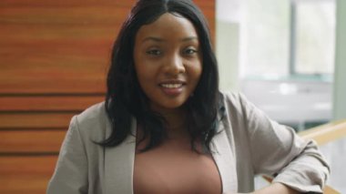 Pozitif Afro-Amerikalı iş kadınının göğüs portresi. Ofiste duruyor, kameraya bakıyor ve gülümsüyor.