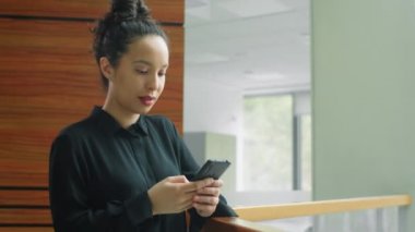 Güzel melez iş kadını ofiste parmaklıklar arkasında duruyor, akıllı telefondan yazı yazıyor ve gülümsüyor.