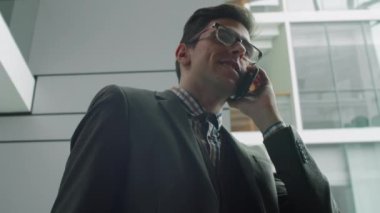 Düşük açı, genç Kafkasyalı iş adamının ofis merkezinde durup cep telefonuyla konuşmasını gösteriyor.