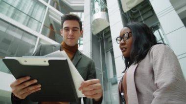 Pozitif beyaz iş adamı ve Afro-Amerikalı iş kadınının, büyük ofis merkezinde dururken dökümanları tartışmalarının düşük açılı görüntüsü