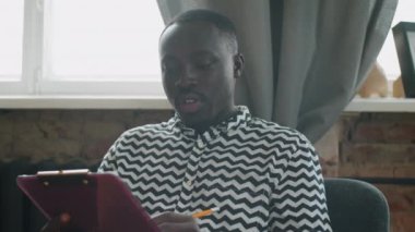 Genç Afro-Amerikalı öğrenci kameraya bakıyor, notlar alıyor ve evdeki çevrimiçi video konuşmalarıyla uzaktan çalışıyor.