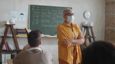 Koruyucu maskeli kadın öğretmen sınıfa giriyor ve covid-19 salgını sırasında çalışırken çok ırklı öğrencilere ders veriyor.