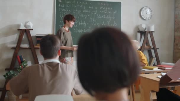 若い女子学生は笑顔で先生や多民族のクラスメートと数学の授業で話しながら 黒板に数式で立っています — ストック動画
