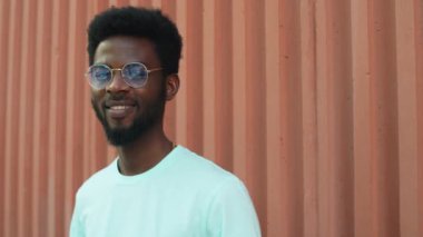Gözlüklü ve günlük tişörtlü genç Afro-Amerikan bir adamın portresi. Kamera önünde gülümseyerek poz verirken yaz günü şehir duvarının yanında dikiliyordu.