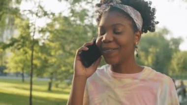 Güneşli yaz gününde parkta yürüyen ve cep telefonuyla konuşan güzel Afro-Amerikalı kadın.