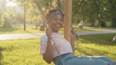Mutlu Afro-Amerikalı kadın güneşli yaz gününde parkta salıncakta sallanırken kameraya bakıp gülümsüyor.