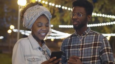 Genç ve pozitif Afro-Amerikalı arkadaşlar akşam parkta yan yana dururken akıllı telefon ekranında gülümsüyor ve bir şeyler tartışıyorlar.