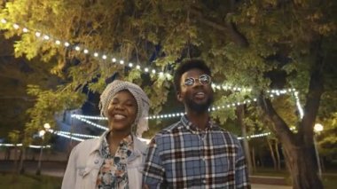Parkta yürürken gülümseyen ve sohbet eden genç Afro-Amerikalı çift akşamları çelenklerle aydınlanıyor.