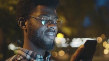 Gözlüklü genç Afro-Amerikalı adamın akıllı telefon ekranına bakarken ve akşam parkta gülümserken yakın plan çekimleri.