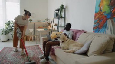 Beyaz bir kadın oturma odasında yerleri süpürüyor ve kanepede dizüstü bilgisayarı ve evcil köpeği olan Afro-Amerikan kocasıyla konuşuyor.