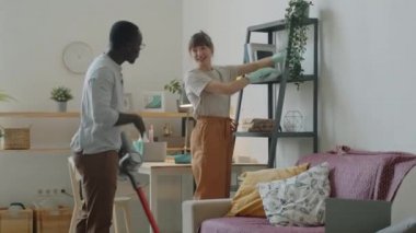 Eldivenli neşeli beyaz kadın kitap raflarını kumaşla temizliyor ve oturma odasını süpürürken Afro-Amerikan kocasıyla sohbet ediyor.