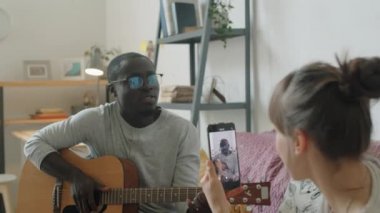 Genç siyahi bir adam gitar çalıp şarkı söylerken Kafkasyalı karısı evde akıllı telefonuyla video kaydediyor.