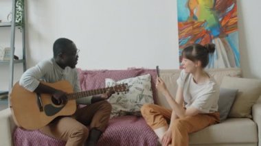 Genç Afrikalı bir adam oturma odasındaki koltukta gitar çalıp şarkı söylerken beyaz karısı akıllı telefonuyla video kaydediyor.