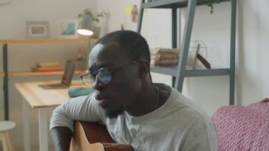 Genç Afro-Amerikalı adam evdeki kanepede oturup gitar çalıyor ve şarkı söylüyor.