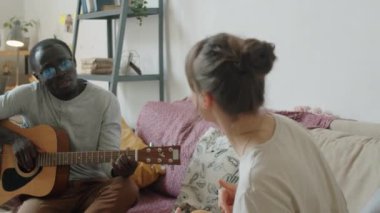 Genç Afrikalı adam evdeki kanepede gitar çalıp şarkı söylerken beyaz karısı müzik ve dansın keyfini çıkarıyor.