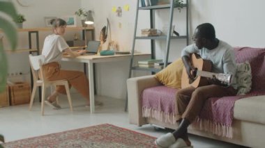 Genç beyaz kadın dizüstü bilgisayar kullanıyor ve oturma odasındaki kanepede gitar çalıp şarkı söylerken Afro-Amerikan kocasını dinliyor.
