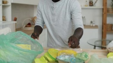 Afro-Amerikalı bir adamın mutfak masasında geri dönüşüm için atıkları düzenlerken plastikleri poşetlere ayırması.