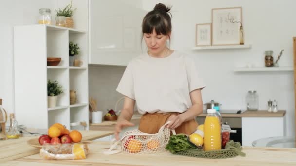 若い白人女性は自宅でキッチンテーブルの上に優しいネットバッグから食料品をアンロード — ストック動画
