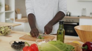 Genç siyah adam, mutfakta sebze salatası hazırlarken ahşap tahtada taze marul kesiyor.