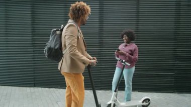 Afro-Amerikalı bir adam şehir caddesinde e-scooter 'a biniyor, kız arkadaşına uğruyor, onu kucaklıyor ve sohbet ediyor.