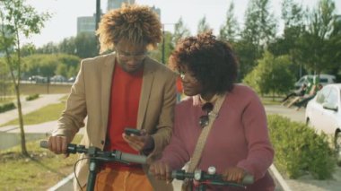Genç Afro-Amerikalı kadın ve erkek e-scooterlarla parkta duruyor, gülümsüyor ve akıllı telefon ekranında bir şeyler tartışıyorlar.