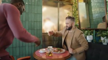 Afro-Amerikalı bir adam beyaz meslektaşını el sıkışarak selamlıyor, kafe masasında oturuyor, fincana çay dolduruyor ve iş yemeğinde onunla konuşuyor.