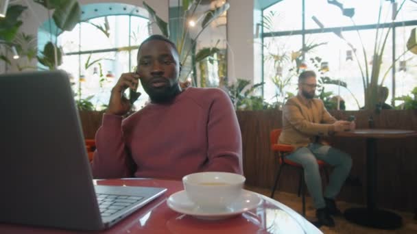 ノートパソコンとティーカップ付きのカフェでテーブルに座っている間に携帯電話で話すカジュアルな服装の黒人実業家 — ストック動画