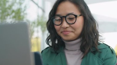 Gözlüklü genç Asyalı iş kadını dışarıda dizüstü bilgisayarla oturuyor ve akıllı telefon kullanıyor.