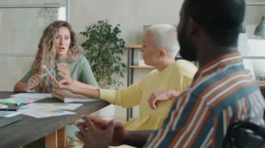 Genç Kafkasyalı iş kadını, ofiste takım toplantısı yaparken tekerlekli sandalyedeki Afro-Amerikalı erkek meslektaşıyla çalışmayı tartışıyor.