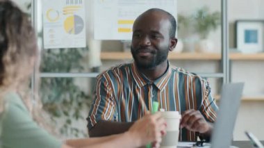 Afro-Amerikalı iş adamı, takım toplantısı sırasında kahve içmek ve beyaz kadın meslektaşıyla konuşmak için ofis masasında oturuyor.
