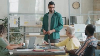 Orta Doğulu işadamı, iki beyaz kadın ve tekerlekli sandalyedeki Afro-Amerikalı adamın önünde duruyor cam ofis duvarındaki kağıtlara bakıyor ve takım toplantısı sırasında sunum yapıyor.