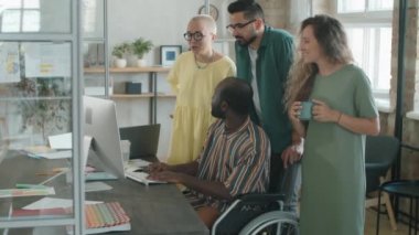 Engelli Afro-Amerikalı iş adamı tekerlekli sandalyede oturuyor ve çok ırklı bir ekiple bilgisayar üzerinde proje tartışıyor.