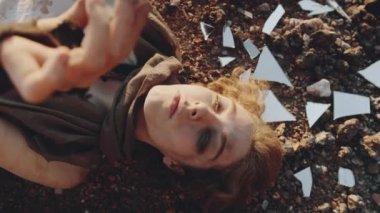 Yüksek açılı, el kameralı, yüzü kirlenmiş genç bir kadının kıyamet sonrası dünyada kırık ayna parçalarının arasında yattığını gösteren bir fotoğraf.