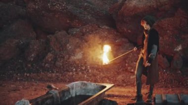 Eski püskü elbiseli genç bir kadın, kıyamet sonrası kamp alanında yürüyor ve kirli küvette suya yanan meşale koyuyor.