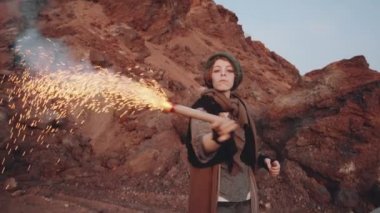 Eski püskü elbiseli genç bir kadın elinde renkli bir sis bombası tutuyor ve kıyamet sonrası dünyadaki kamp alanında vinil müzik eşliğinde dans ediyor.