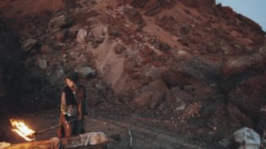 Yaşlı elbiseli bir kadının elinde yanan bir meşale tutarken ve distopya dünyasında kirli suyla dolu paslı küvetin yanında kameraya bakarken çekilen görüntüsü.