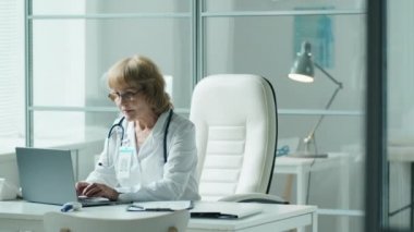 Laboratuvar önlüklü kıdemli bayan doktor laptopta çalışıyor ve tıp bürosundaki masada notlar alıyor.