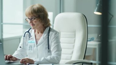 Laboratuvar önlüklü yaşlı kadın doktor portresi. Steteskop boynunda. Masasında oturup kameraya gülümseyerek poz veriyor.