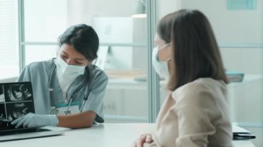 Koruyucu maskeli İspanyol kadın doktor ve eldivenli kadın doğum öncesi konsültasyon sırasında laptoptaki fetal ultrason taramasını beyaz hamile kadına açıklar.