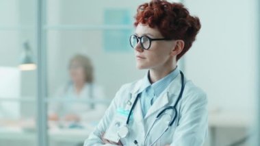 Kısa kızıl saçlı, kolları çapraz duran ve klinikte kameraya poz veren kendine güvenen genç bir kadın doktorun portresi.