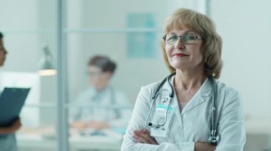 Laboratuvar önlüklü yaşlı kadın doktor portresi. Kolları çapraz duruyor ve poz veriyor. Klinikte gülümsüyor.