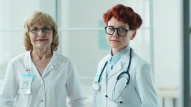 Laboratuvar önlüklü yaşlı ve genç kadın doktorların portresi. Klinikte yan yana durup kameraya bakıyorlar.