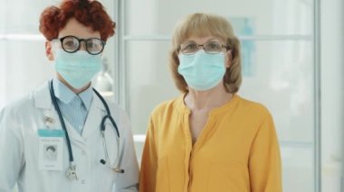 Korunaklı maskeli genç kadın doktor ve yaşlı kadının portresi klinikte yan yana duruyor ve Coronavirus salgını sırasında kameraya poz veriyor.