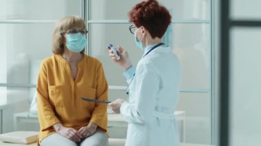 Maskeli kadın doktor, kızılötesi termometresi olan ve klinikte covid-19 salgını sırasında sağlık kontrolü sırasında panoya sıcaklık ölçümleri yazan yaşlı bir kadının alnını tarıyor.