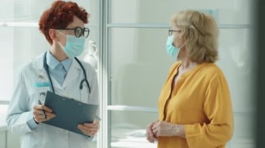 Maskeli genç bayan doktor panoya yazı yazıp klinikte covid-19 salgınında sağlık danışmanlığı yaparken yaşlı kadınla konuşuyor.